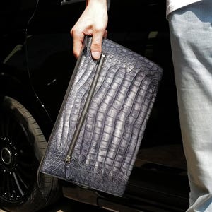 May include: A rectangular clutch bag featuring a dark gray crocodile skin texture with a prominent vertical zipper pocket on the front. The accessory has a structured design with a metallic zipper pull and a consistent scale pattern throughout.