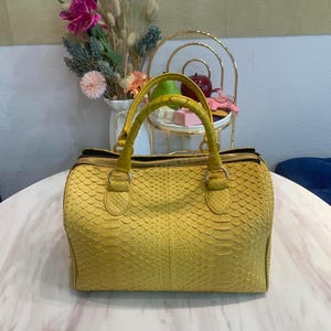 May include: A bright yellow handbag with a textured snakeskin-like pattern, featuring two sturdy top handles, a rectangular base, and a gold-toned zipper closure along the top edge. The bag has a structured shape with visible stitching details.