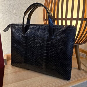 May include: A black handbag with a textured snakeskin pattern and two top handles. The bag features a rectangular shape with a zippered top closure. The word Sandyakala is printed in a subtle font across the center of the front panel.