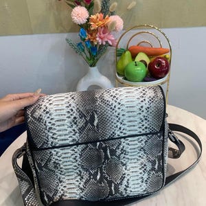 Peut inclure: Un sac à bandoulière rectangulaire présentant un motif texturé en peau de serpent dans des tons de blanc, gris et noir. Le sac possède un rabat, un passepoil noir le long des bords et une longue bandoulière noire réglable sur les côtés.