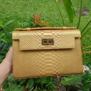 Peut inclure: Un sac à main doré structuré avec un motif texturé en peau de serpent, un rabat rectangulaire et un fermoir tournant en métal doré au centre. Le sac possède une poignée supérieure et une silhouette rigide et carrée avec une finition métallique.
