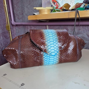 May include: A rectangular clutch bag featuring a textured, scale-like pattern in deep brown with a prominent, vertical bright blue stripe running down the centre. The bag has a folded flap closure and is positioned near a ruler showing metric markings.