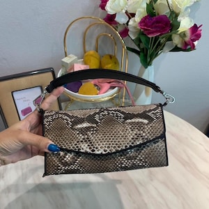 May include: A rectangular clutch bag featuring a brown and black snakeskin pattern with a black trim and a detachable black handle. The bag has a fold-over flap closure with silver-toned metal hardware clips on each side for the handle attachment.