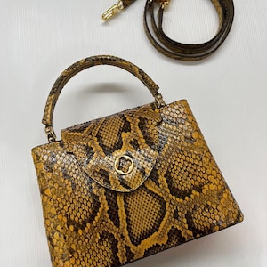 May include: A structured handbag featuring a yellow and dark brown snakeskin pattern. The bag has a single top handle, a flap closure with a circular gold-toned metal emblem, and a detachable, adjustable shoulder strap with gold-toned hardware.