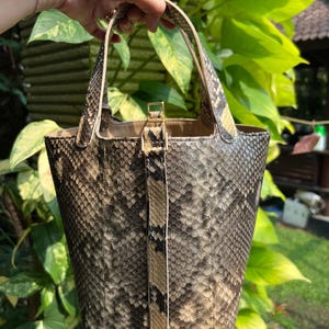 Puede incluir: Un bolso tipo cubo con un patrón de piel de serpiente texturizado en tonos marrón, gris y crema. El bolso tiene dos asas superiores y una correa de cierre vertical en el centro con una hebilla y herrajes de metal dorado, de diseño estructurado.