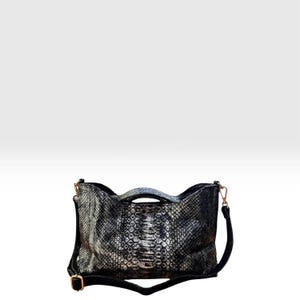 Könnte beinhalten: Eine rechteckige Handtasche mit einer metallisch silbernen und schwarzen Schlangenledermuster-Textur. Die Tasche verfügt über einen Tragegriff, einen abnehmbaren schwarzen Schulterriemen mit goldfarbenen Metallbeschlägen und eine strukturierte Form.