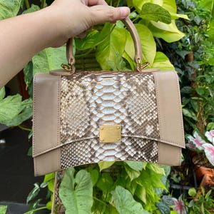 Peut inclure: Un sac à main structuré doté d'un panneau central au motif peau de serpent texturé dans des tons marron, blanc et beige, flanqué de panneaux latéraux en cuir beige lisse, d'une poignée supérieure et d'un fermoir métallique doré.