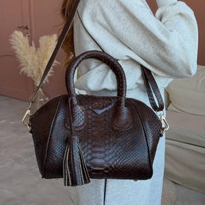 Könnte beinhalten: Eine dunkelbraune Handtasche mit strukturiertem Schlangenledermuster, ausgestattet mit zwei abgerundeten Tragegriffen, einem abnehmbaren Schulterriemen und einer dekorativen Quaste an einem Griff. Die Tasche hat eine strukturierte Form mit goldfarbenen Metallbeschlägen.