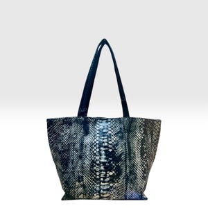 Pode incluir: Uma bolsa tote com um padrão de pele de cobra em preto e prateado com acabamento texturizado. A bolsa possui duas alças de ombro longas e pretas fixadas no topo, uma base retangular larga e uma silhueta que se alarga para cima.