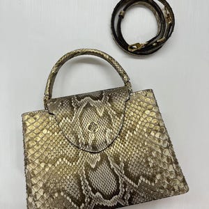 May include: A structured handbag featuring a metallic gold and brown snakeskin pattern. The bag has a single top handle, a flap closure with a circular gold-toned emblem, and a detachable, coiled shoulder strap with gold-toned hardware.