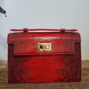 Könnte beinhalten: Eine strukturierte rote Handtasche mit einer texturierten Eidechsenhaut-Optik. Die Tasche verfügt über einen Tragegriff und einen rechteckigen Drehverschluss aus goldfarbenem Metall an der Frontklappe. Das Design umfasst eine horizontale Klappe.