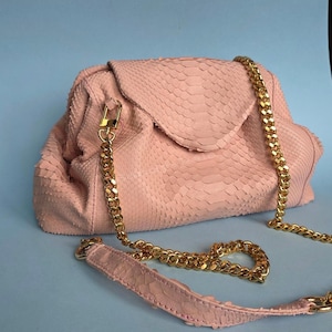 May include: A light pink handbag with a textured snakeskin pattern, featuring a structured frame, a fold-over flap, and a thick gold-tone metal chain strap. The strap includes a matching pink snakeskin leather shoulder pad and gold metal clasps.