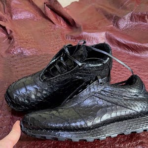 May include: A pair of black trainers featuring a textured, scale-like pattern across the upper. The shoes have black laces, a low-top silhouette, and a thick, lugged black rubber sole. The interior lining appears to be dark and fuzzy.