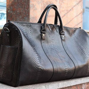 Può includere: Un borsone nero con un motivo testurizzato in pelle di serpente. La borsa include due manici superiori, una chiusura superiore con cerniera e una tasca laterale. È dotata di una base strutturata e di attacchi in metallo per una tracolla.