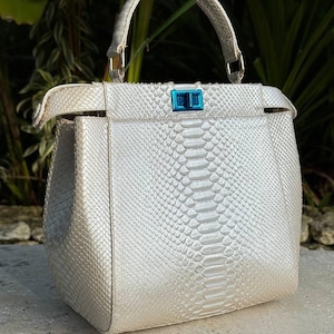 Puede incluir: Un bolso estructurado con una textura de piel de serpiente en color blanco roto brillante. El bolso incluye un asa superior, una estructura rectangular y un llamativo cierre de giro en color azul metálico centrado en la solapa frontal.