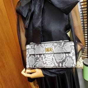 Peut inclure: Un sac à main rectangulaire présentant un motif peau de serpent gris et blanc avec une finition texturée en écailles. Le sac comprend une poignée supérieure, un rabat et un fermoir tournant en métal doré poli centré sur le panneau avant.