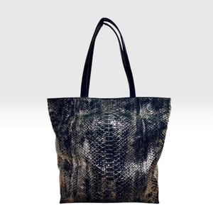 May include: A tote bag featuring a black and metallic silver snakeskin pattern. The bag has a rectangular body with a wide base and two tall, thin black shoulder straps attached to the top edge. The surface texture mimics reptile scales.