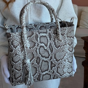 Peut inclure: Un sac à main structuré présentant un motif peau de serpent marron et blanc. Le sac comprend deux poignées tressées et un pompon décoratif suspendu sur un côté. Il a une forme rectangulaire avec des anneaux métalliques argentés sur les côtés.