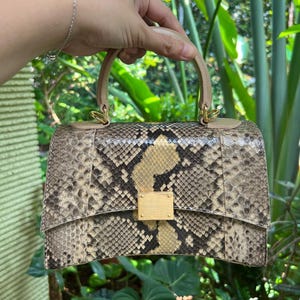 Peut inclure: Un sac à main structuré avec un motif peau de serpent dans des tons beige, marron et noir. Le sac présente un bord inférieur incurvé, une poignée supérieure unique de couleur beige clair et un fermoir métallique doré rectangulaire.