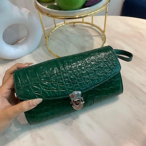 Può includere: Una pochette verde scuro con una texture lucida in pelle di coccodrillo e una chiusura a pressione in metallo color argento. La borsa rettangolare presenta un design a patta e un piccolo cinturino laterale, con un aspetto strutturato.