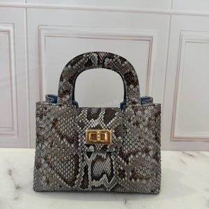 Peut inclure: Un sac à main structuré présentant un motif peau de serpent gris, noir et blanc. Le sac possède une poignée supérieure unique, un rabat rectangulaire fermé par un fermoir pivotant en métal doré et une doublure intérieure bleue visible.