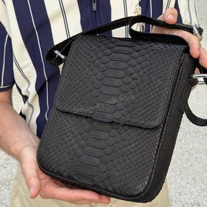 Puede incluir: Un bolso tipo mensajero negro con un patrón de piel de serpiente texturizado en todo el exterior. El bolso tiene un cierre de solapa rectangular con un logotipo sutilmente grabado que dice sandyakala. Incluye una correa de hombro negra ajustable.