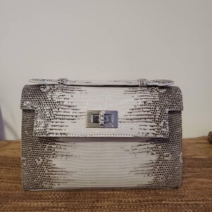 Pode incluir: Uma mala de mão estruturada com um padrão texturizado inspirado em répteis em tons de branco, cinzento e preto. A mala inclui uma pega superior, um fecho de aba retangular e um mecanismo de fecho giratório em metal prateado.