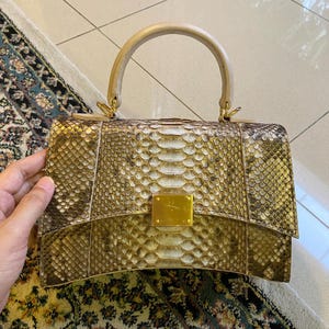 Peut inclure: Un sac à main structuré présentant un motif texturé en peau de serpent dans des tons dorés, marron et crème. Le sac possède une poignée supérieure incurvée, un fermoir métallique doré rectangulaire sur le rabat et une base arquée.