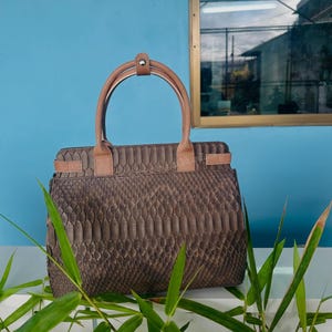 May include: A structured handbag featuring a dark brown, textured snakeskin-patterned exterior. The bag includes two light tan leather handles secured with a central snap tab, and matching tan leather side straps with metallic hardware accents.