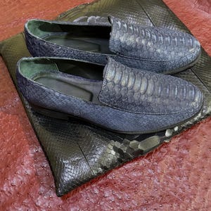 May include: A pair of blue slip-on loafers featuring a textured snakeskin pattern on the exterior. The shoes have a rounded toe, a low profile, and a dark interior lining. The loafers are positioned on a dark, textured, rectangular surface.