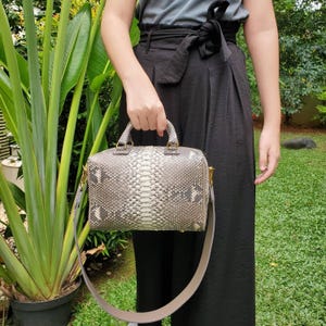 Peut inclure: Un sac à main structuré présentant un motif peau de serpent gris et blanc avec une finition texturée en écailles. Le sac comprend deux anses courtes, des attaches en métal doré et une longue bandoulière amovible gris clair lisse.