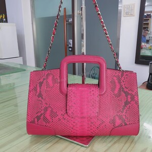 May include: A vibrant pink handbag featuring a textured snakeskin pattern, a structured rectangular shape, a central top handle, and a long silver chain shoulder strap. The bag includes a flap closure and solid pink leather-like corner accents.