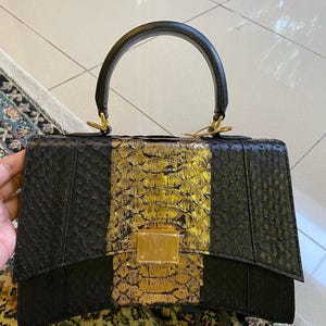 May include: A structured handbag featuring a textured black snakeskin pattern on the sides and a metallic gold snakeskin panel in the centre. The bag includes a single black top handle with gold hardware and a rectangular gold-toned metal clasp.