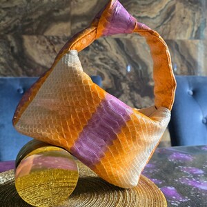Könnte beinhalten: Eine kleine Handtasche mit einem strukturierten, schlangenhautartigen Muster in abwechselnden Streifen aus leuchtendem Orange, sanftem Violett und Off-White. Die Tasche hat einen integrierten Griff, der durch einen Knoten oben geformt ist.
