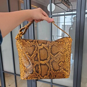 May include: A mustard yellow and black snakeskin patterned handbag with a rectangular shape, a top zip closure, and a single shoulder strap attached with a knotted loop detail on one side, featuring a textured, scaled surface throughout.