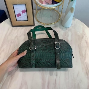 Può includere: Una borsa a mano verde scuro con motivo testurizzato in pelle di serpente, caratterizzata da una parte superiore arrotondata, un dettaglio con fibbia argentata sul davanti e due manici verdi abbinati. La borsa ha una chiusura a zip e forma.