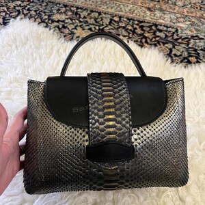 May include: A structured handbag featuring a metallic silver and black snakeskin-textured finish. The bag has a solid black leather flap, a single top handle, and a central vertical panel with a black leather tab closure on the front exterior.