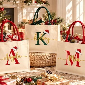Può includere: Tre borse tote in tela color bianco sporco con manici rossi o verdi. Ogni borsa presenta una grande lettera iniziale in rosso o verde, un nome in corsivo dorato e una piccola grafica di un cappello di Babbo Natale.