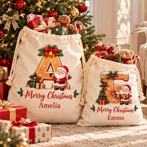Può includere: Due sacchi regalo in tela color crema con coulisse, caratterizzati da lettere in stile pan di zenzero A ed E, decorati con figure di Babbo Natale, agrifoglio e la scritta Merry Christmas con i nomi Amelia ed Emma in rosso.