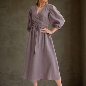 May include: A mauve midi-length wrap dress featuring a V-neckline, voluminous three-quarter length puff sleeves with gathered cuffs, and a matching fabric tie belt at the waist. The dress has a full skirt and is made of a textured fabric.