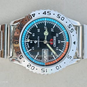 Op de afbeelding: Een roestvrijstalen Seiko 5 automatisch polshorloge met een zwarte wijzerplaat voorzien van 24-uurs markeringen, een dag-datumvenster en lichtgevende wijzers. De draaibare ring is zilverkleurig met zwarte cijfers en een blauwe en oranje rand.