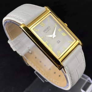 May include: A rectangular Seiko Quartz wristwatch featuring a gold-tone case and a white dial with gold Roman numeral hour markers. The watch has a white leather strap with a textured crocodile-embossed pattern and gold-tone hands.