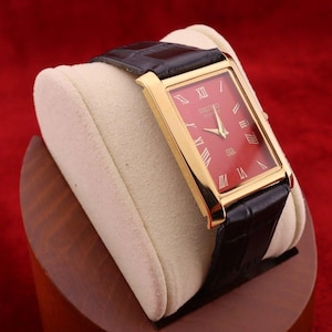 May include: A rectangular wristwatch featuring a polished gold-tone case and a vibrant red dial with gold Roman numeral hour markers. The watch is equipped with a black leather strap that has a textured, embossed finish and a gold-tone buckle.