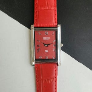 Könnte beinhalten: Eine rechteckige Seiko Quartz Armbanduhr mit einem leuchtend roten Zifferblatt, silberfarbenen römischen Ziffern als Stundenmarkierungen und passenden Zeigern. Die Uhr hat ein strukturiertes rotes Lederarmband mit Krokodilprägung und ein silberfarbenes Gehäuse.