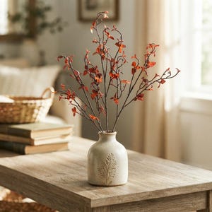 May include: A small, speckled off-white ceramic vase with a narrow neck and a debossed fern leaf pattern on the front. The vase holds several thin, dark brown branches adorned with small, vibrant orange and red leaves extending upwards.