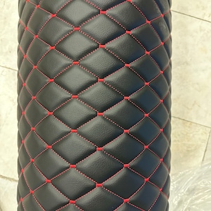 Puede incluir: Un rollo cilíndrico negro con un patrón de diamantes acolchado y costuras rojas vibrantes. La superficie tiene un acabado texturizado y el diseño geométrico repetitivo cubre todo el exterior visible del objeto en una cuadrícula.