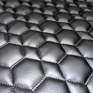 May include: A black material featuring a repeating hexagonal quilted pattern with raised, padded sections. The surface has a textured, leather-like appearance with visible stitching defining each individual hexagon shape across the entire piece.
