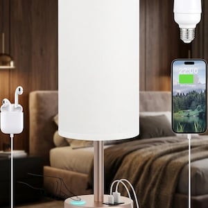 May include: A cylindrical white fabric lampshade sits atop a metallic rose gold base featuring a touch-sensitive power button, two USB charging ports, and a power cord. The lamp is displayed with a white LED bulb, a smartphone, and wireless earbuds.