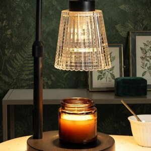 May include: A black metal candle warmer lamp featuring a clear, ribbed glass shade and a circular base. The lamp is positioned over a brown glass jar candle that is illuminated from above, casting a warm glow on the surface below.