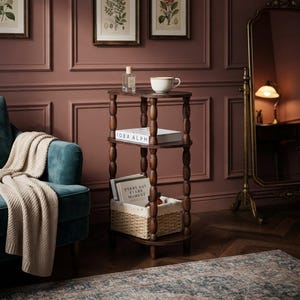 May include: A three-tiered dark wood side table with turned spindle legs. The top shelf holds a clear glass bottle and a white ceramic mug. The middle shelf contains a book titled IDEA ALPH. The bottom shelf holds a woven basket with books.